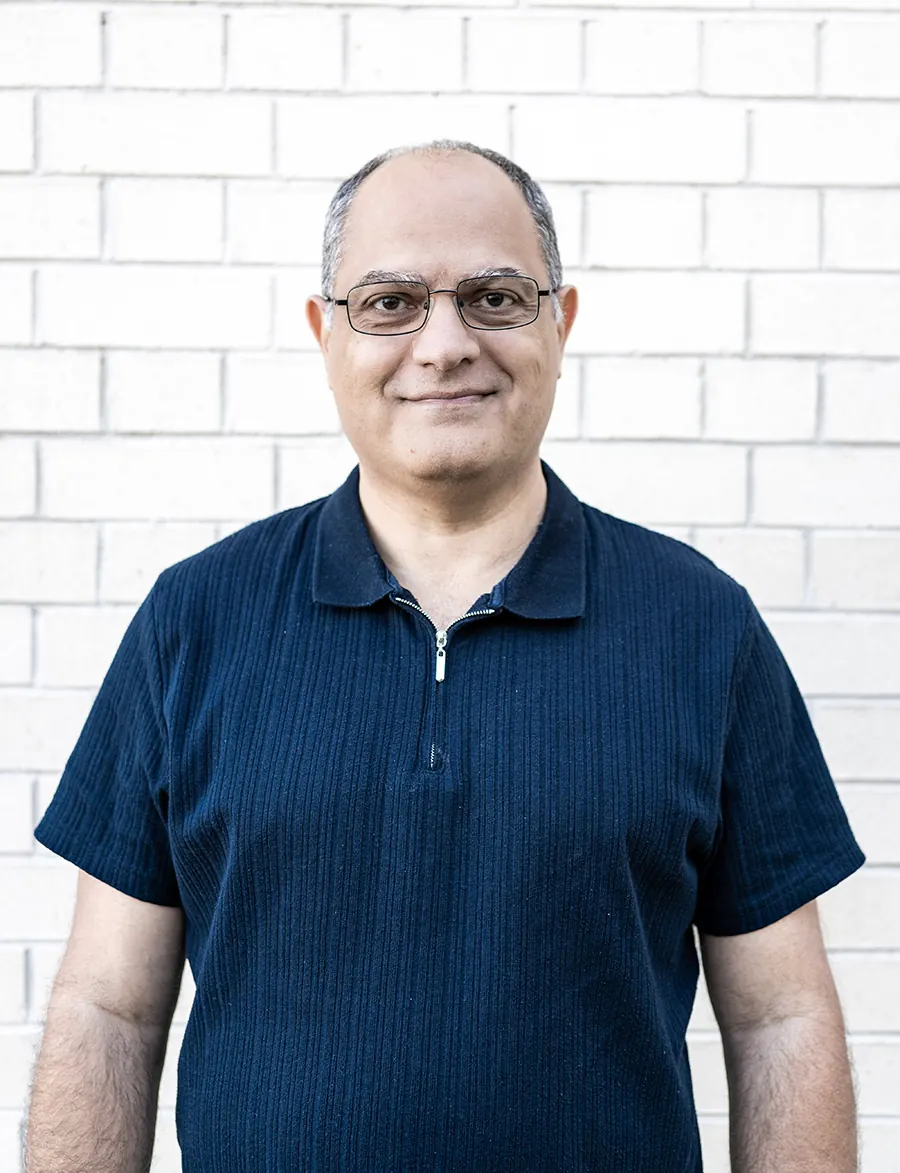 Sam George, wearing glasses and a navy polo shirt, in front of a white brick wall